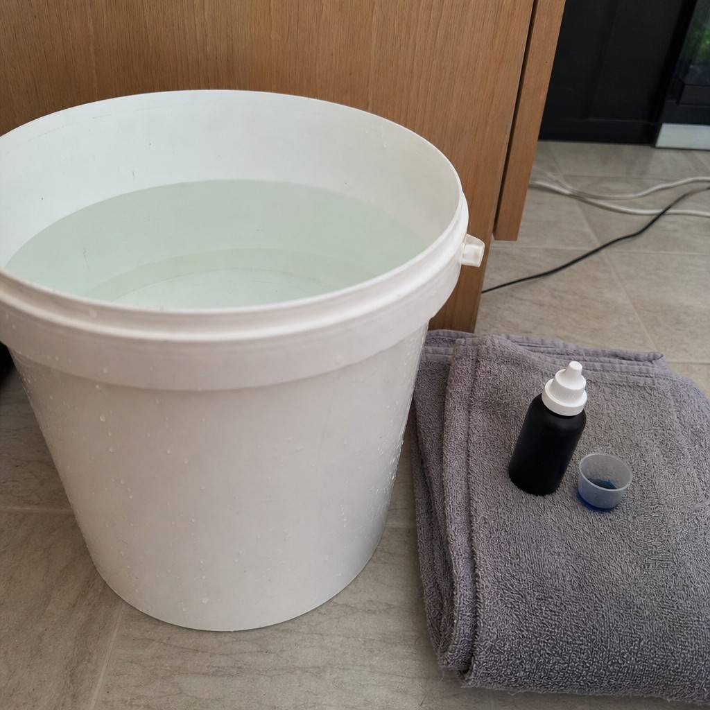 A small bottle of water conditioner and a dosing cap next to a filled bucket on a kitchen floor
